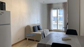Una sala de estar moderna y luminosa, con un sofá, una mesita auxiliar y una mesa de comedor con sillas. Se observa un refrigerador a la izquierda, y una puerta corrediza de vidrio da a un balcón.