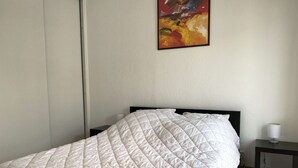 Una cama bien hecha con colcha blanca, una mesita de noche con lámpara y un cuadro en la pared.