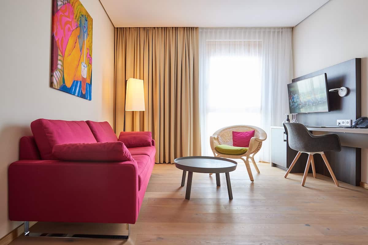 A modern hotel room with a red sofa, a round table, a wicker chair, a desk with a chair, a TV, and a painting on the wall.