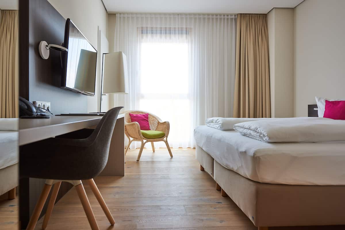 A hotel room with a bed, a desk with a chair, a television, and a window with curtains.