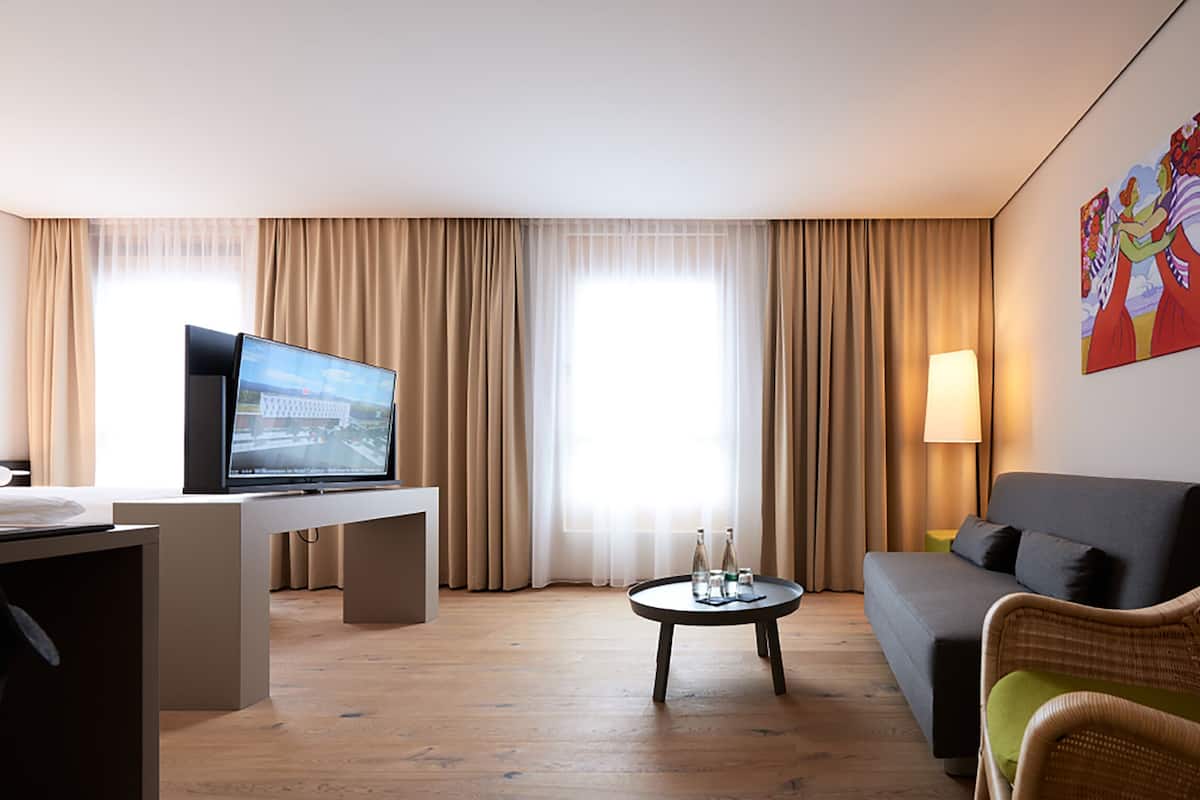A modern hotel room with a flat-screen TV on a desk, a sofa, a small round table, and a large window with curtains.