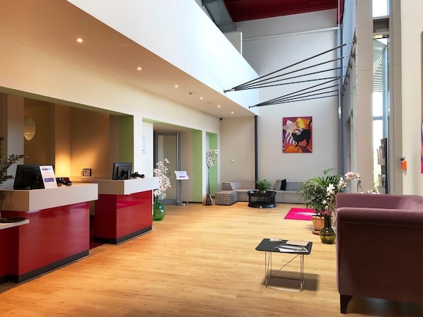 A modern reception area with a wooden floor, red reception desks, and a seating area with a sofa and chairs.