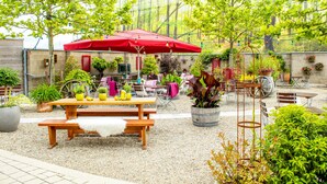 Un espace extérieur aménagé en terrasse, avec une table et des bancs en bois, entouré de plantes en pot et agrémenté d’un parasol rouge.