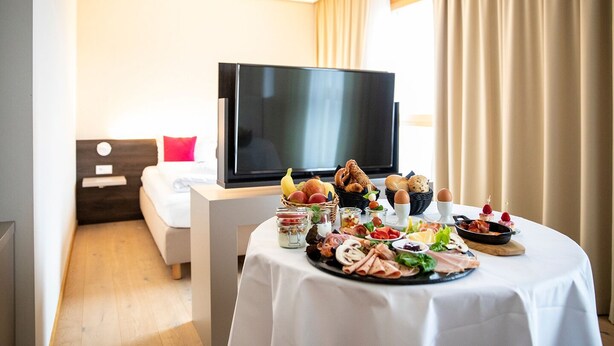 Une chambre d’hôtel avec une table à manger dressée, sur laquelle sont disposés divers aliments, un lit, une télévision et une fenêtre avec des rideaux.
