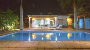 Una piscina con suelo de baldosas, un patio techado con mobiliario exterior y palmeras.