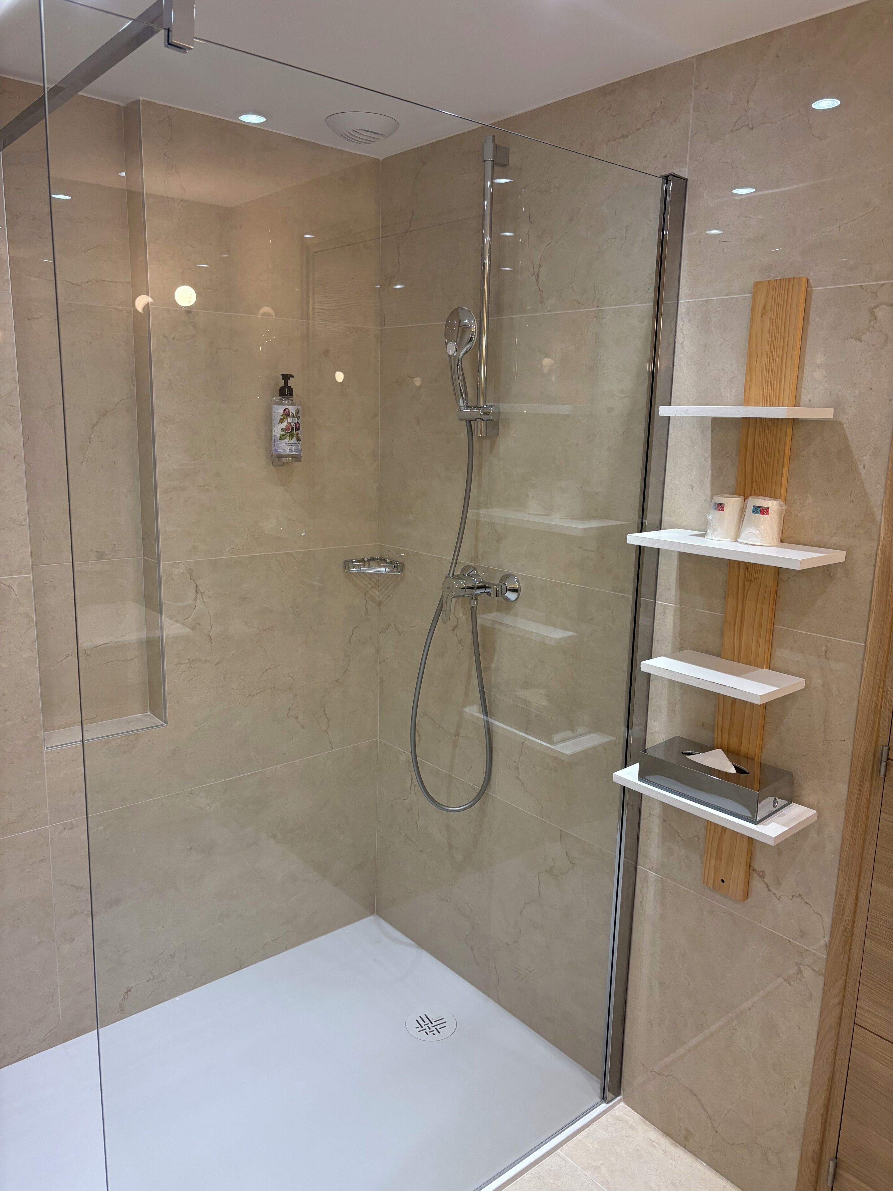 Eine begegnete Dusche mit Handbrause, am Wand montiertem Wasserhahn und Seifenspender. Holzregale mit Pflegeprodukten sind vorhanden.
