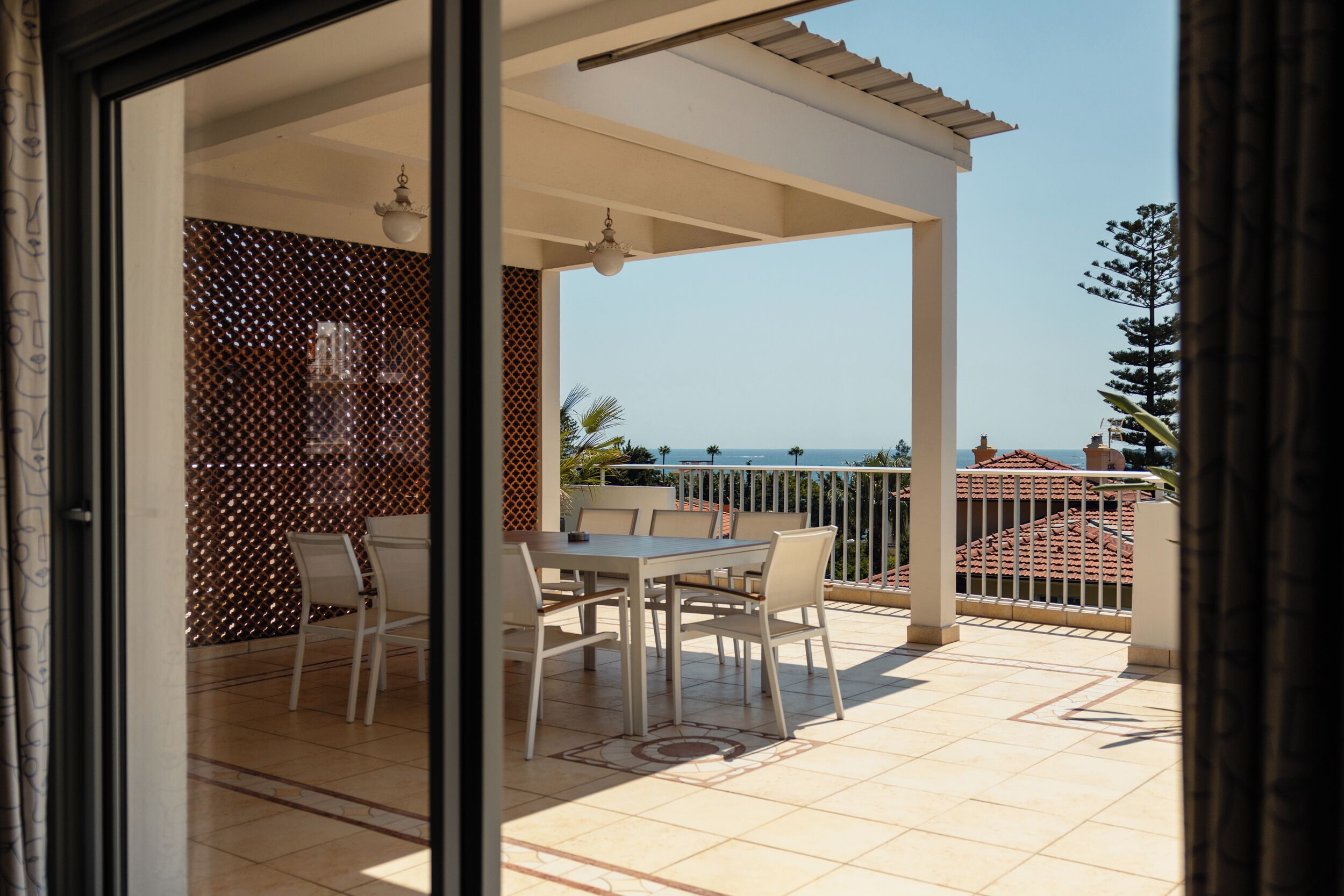 A balcony with a table and chairs, a view of the sea, and a building with red-tiled roofs in the background.