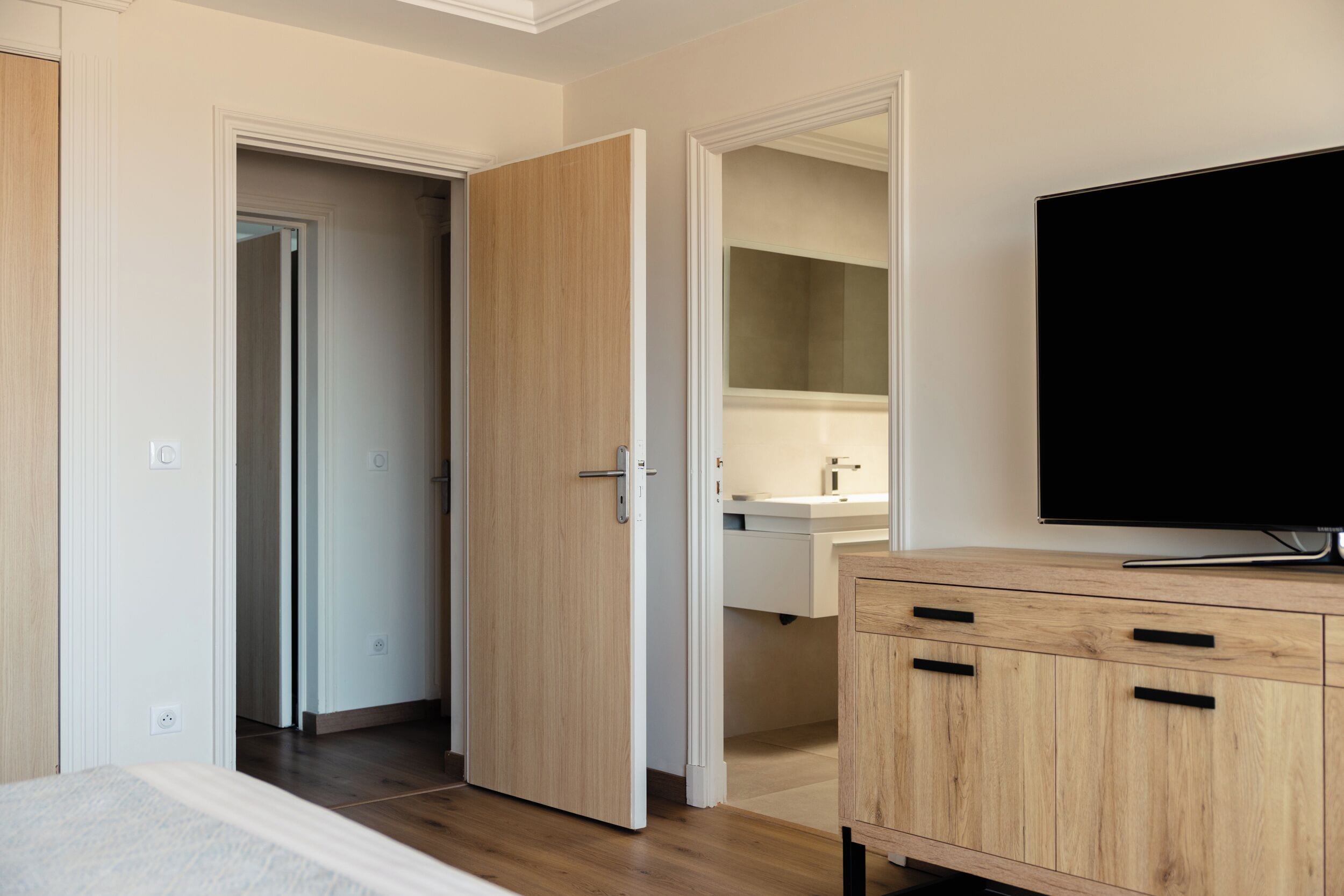A hotel room with a wooden wardrobe, a television, and a bathroom with a mirror and sink.