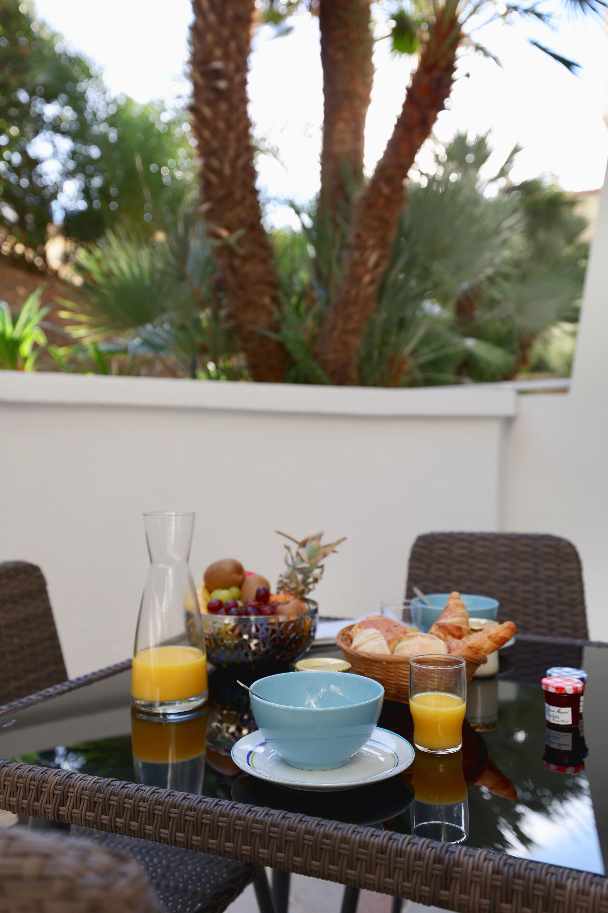 A breakfast table with a glass of orange juice, a bowl of fruit, croissants, and a bottle of jam.