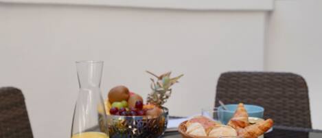 A breakfast table with a glass of orange juice, a bowl of fruit, croissants, and a bottle of jam.