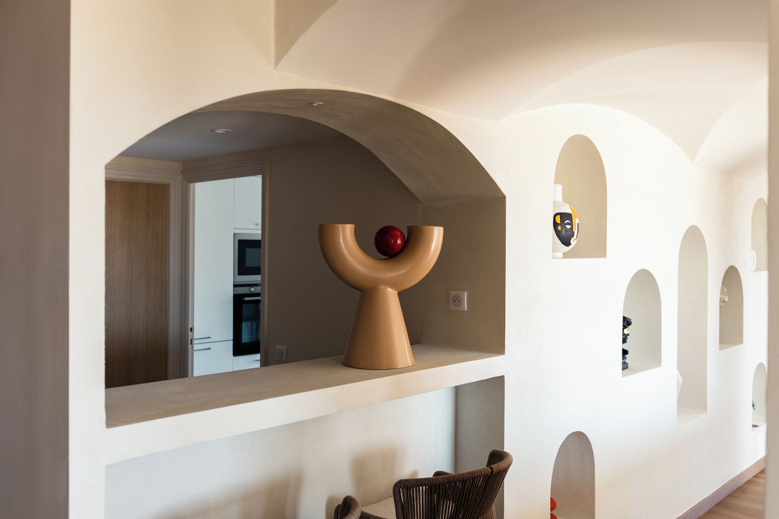 A modern interior with a kitchen area, a unique sculptural piece on a shelf, and a seating area with a woven chair.