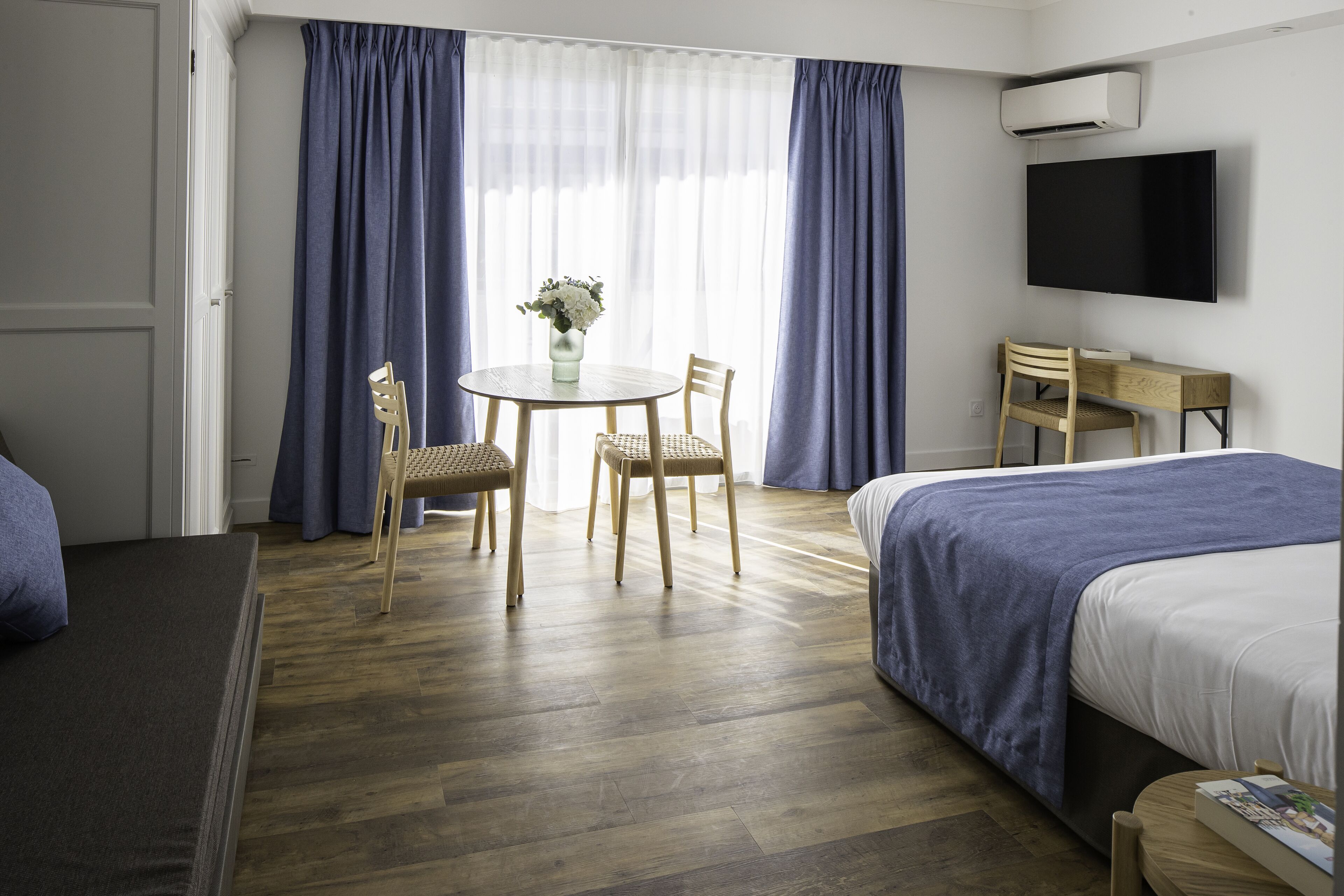 A hotel room with a bed, a desk, a chair, a table with a vase of flowers, and a TV mounted on the wall.