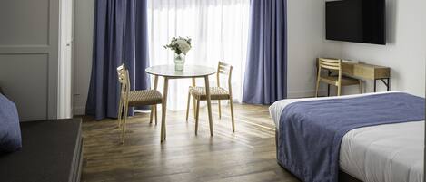 A hotel room with a bed, a desk, a chair, a table with a vase of flowers, and a TV mounted on the wall.