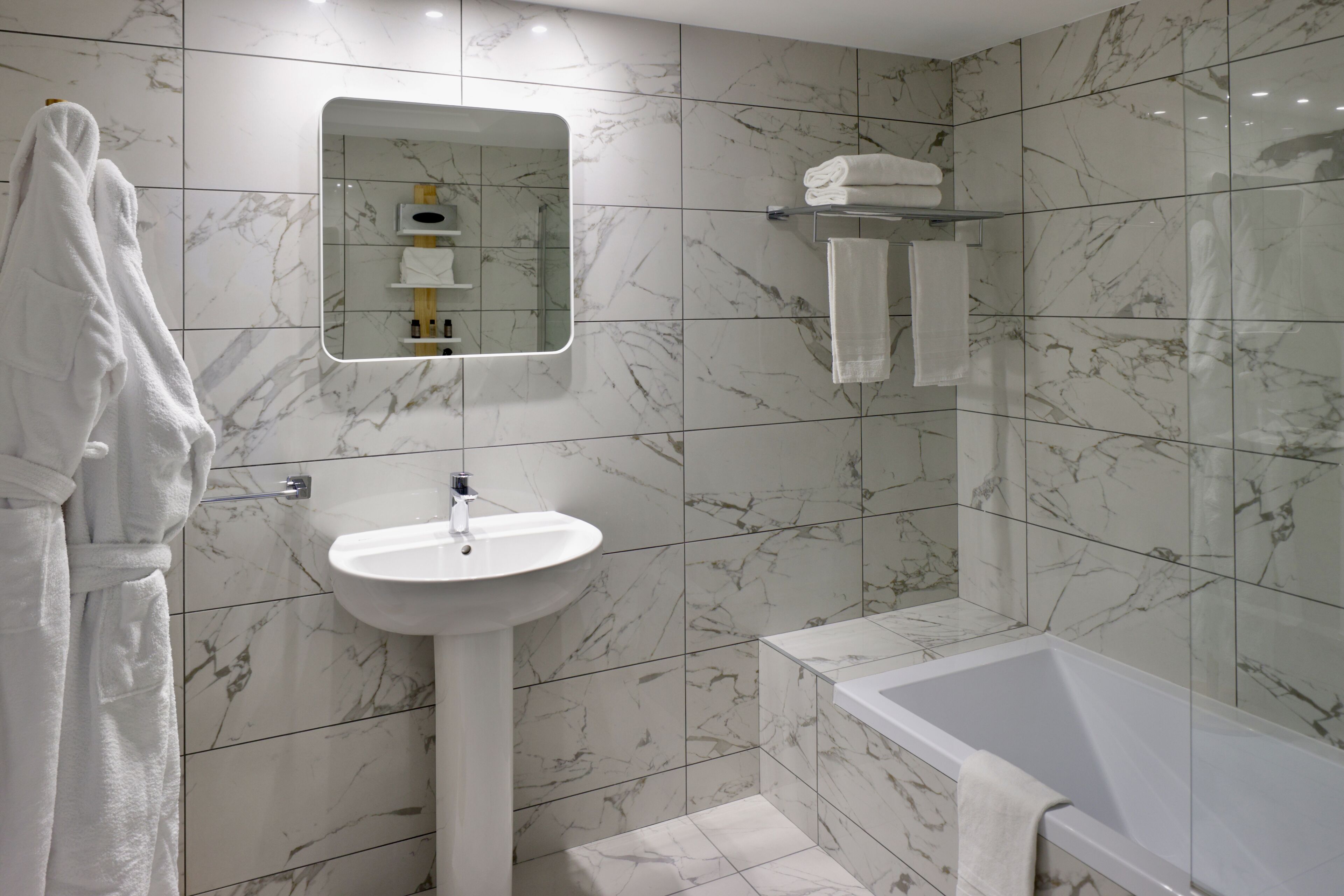 A modern bathroom with a marble-tiled wall, a pedestal sink, a bathtub, and a mirror above the sink.