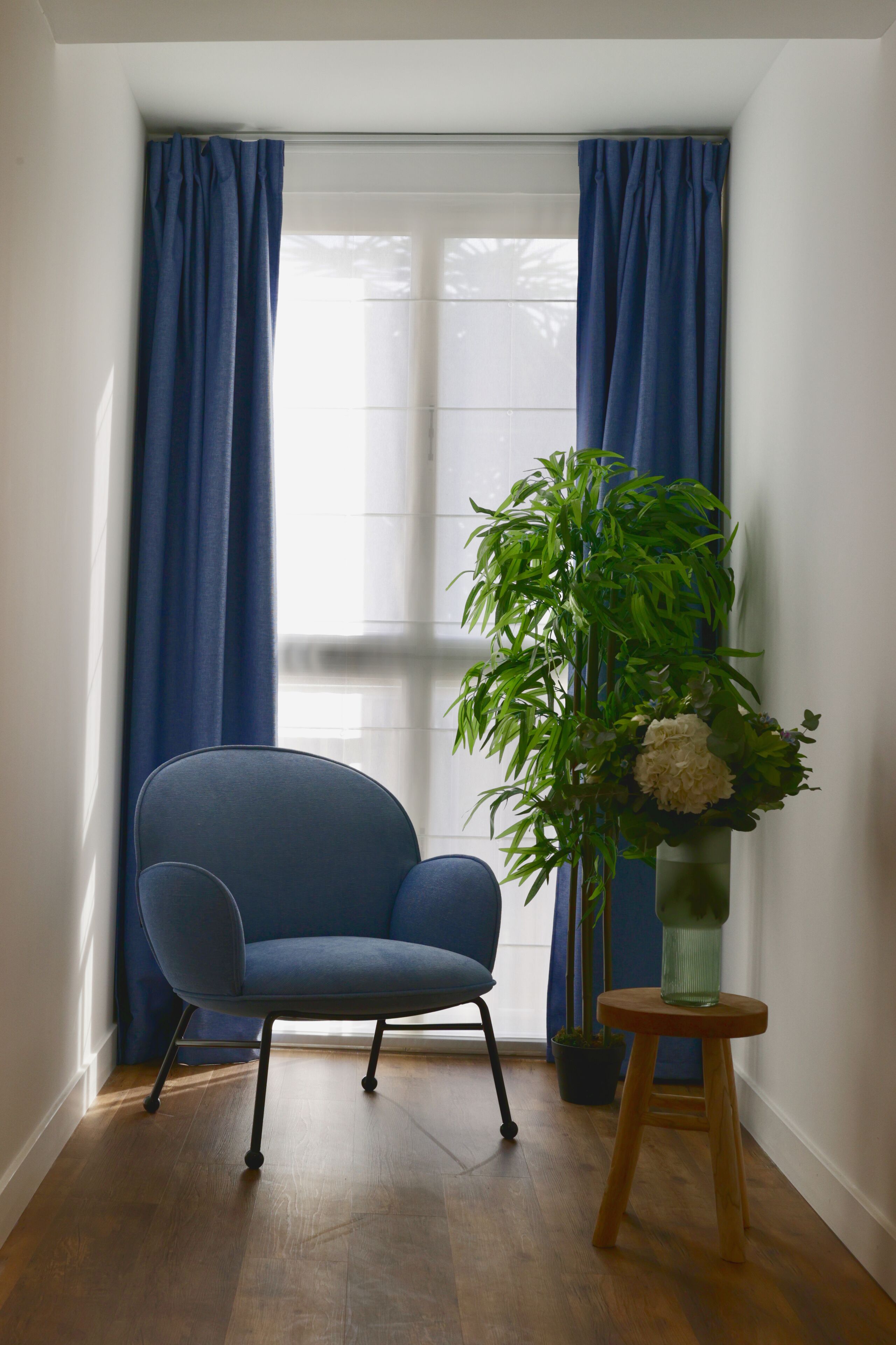 A room with a blue chair, a potted plant, and a vase on a small wooden stool.