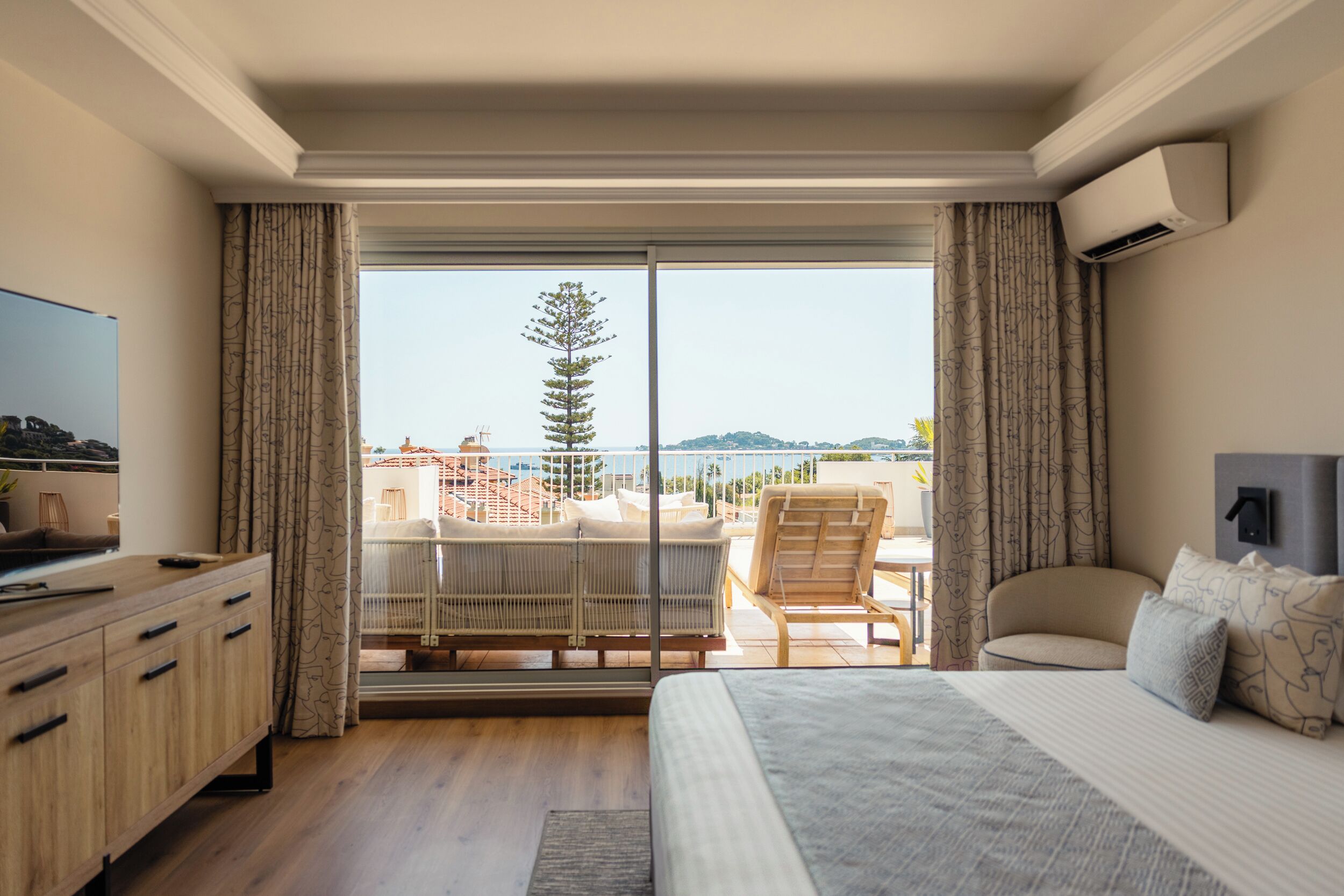 A hotel room with a balcony, a bed, a desk, and a view of the sea and hills.