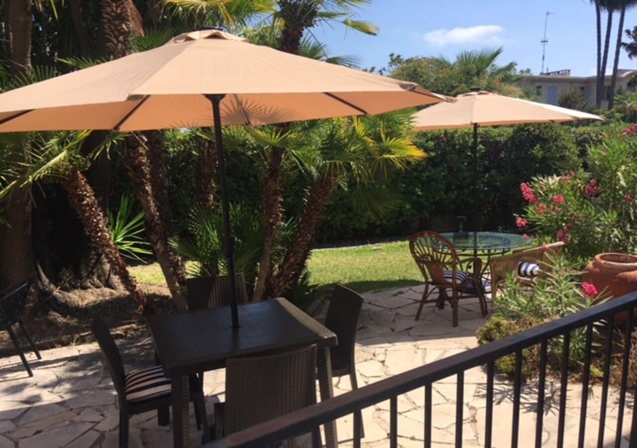 Un espace patio extérieur avec une table et des chaises sous un grand parasol, entouré d’une végétation luxuriante et d’une petite piscine.