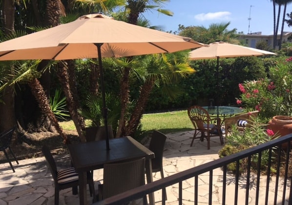 An outdoor patio area with a table and chairs under a large umbrella, surrounded by lush greenery and a small pool.