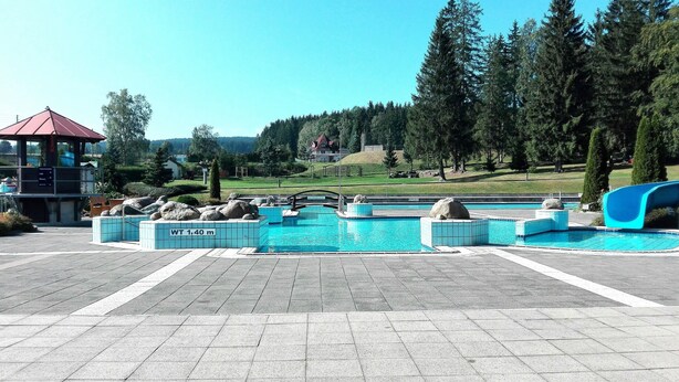 A swimming pool with a water slide, surrounded by a paved area with large rocks and a small building with a red roof. - Vacation home with covered terrace No pets  (Friedrichroda)