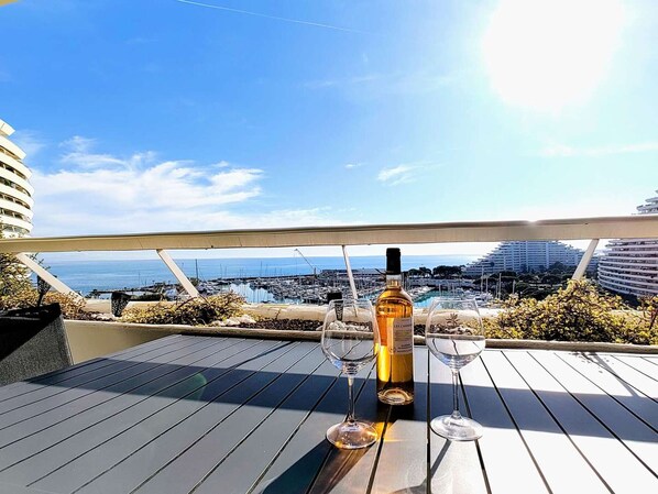 A terrace with a bottle of wine and two glasses, overlooking a marina and buildings.