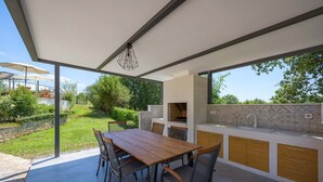 An outdoor kitchen with a dining table and chairs, a built-in grill, and a view of the garden. - Villa Eufemia (Porec)