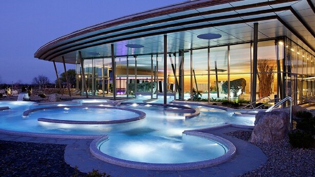 Un edificio moderno con fachada de vidrio, zona de piscina con múltiples jacuzzis y asientos al aire libre.