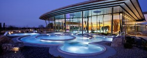 Un edificio moderno con fachada de vidrio, zona de piscina con múltiples jacuzzis y asientos al aire libre.