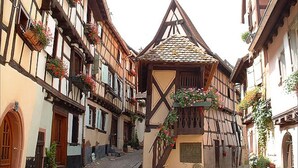 Una estrecha calle empedrada bordeada por edificios tradicionales con entramado de madera, con jardineras llenas de flores y una pequeña construcción de piedra en el centro.