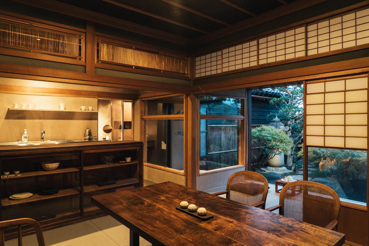 A traditional Japanese-style kitchen with wooden cabinets, a dining table, and a view of a garden through a sliding door.