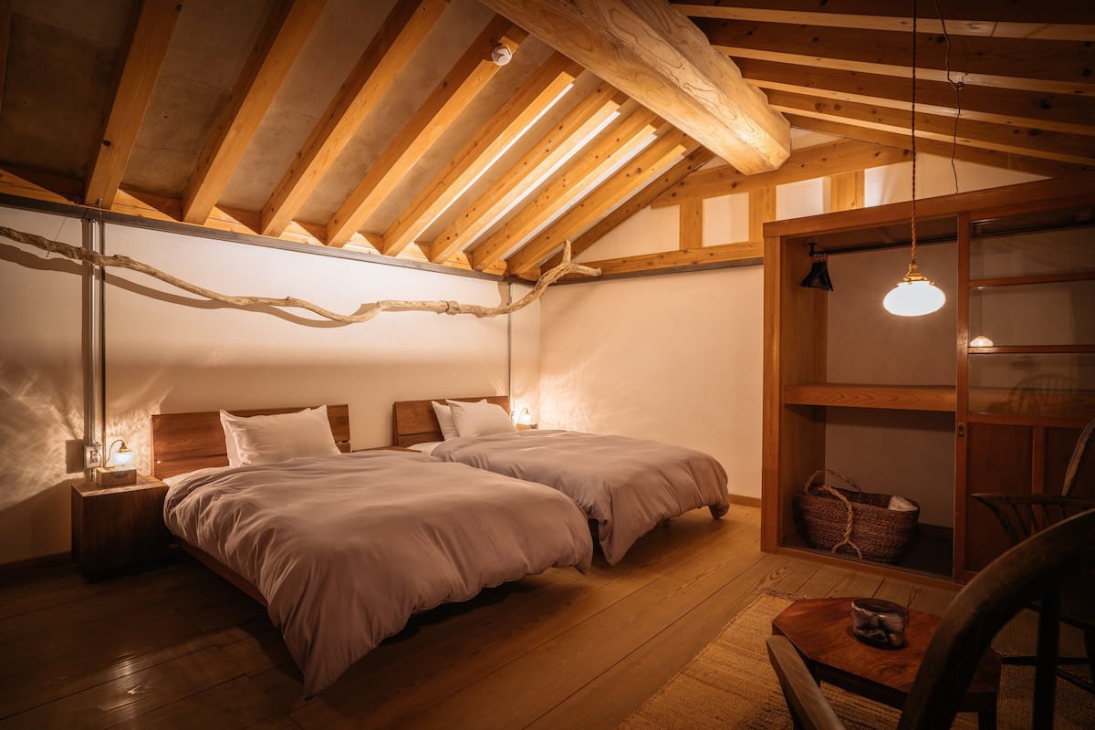 A cozy bedroom with two beds, wooden beams, a hanging light fixture, and a small table with a basket.