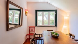 Un coin repas avec une table en bois, des chaises, un fauteuil rouge, un saladier de fruits, un miroir et une fenêtre donnant sur un paysage verdoyant.