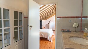 Un dormitorio con techo de madera, una cama con colcha blanca y un baño con lavamanos y espejo.