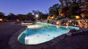 Une piscine avec un mur en pierre et un système d’éclairage, entourée d’arbres et bordée d’une terrasse en briques.