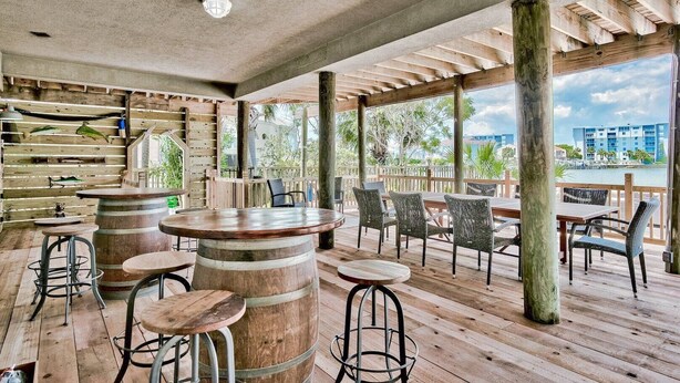 Eine Terrasse im Freien mit Holztischen und -stühlen, ein Barbereich mit Fässern und Blick auf das Meer und Gebäude.