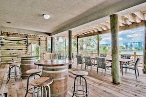 An outdoor patio with wooden tables and chairs, a bar area with barrels, and a view of the ocean and buildings. - Harborfront Home with Pool, Dock & Tiki Cruise (Destin)