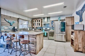 A modern kitchen with a central island, bar stools, stainless steel appliances, and decorative wall art. - Harborfront Home with Pool, Dock & Tiki Cruise (Destin)