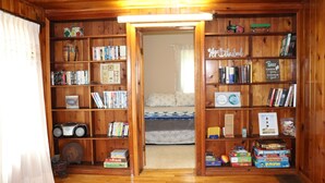 A cozy room with wooden walls and a bookshelf filled with books and games. A bed with a blue and white patterned bedspread is visible in the background.