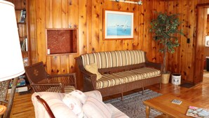 A cozy room with wooden walls, a wicker sofa, a cushioned chair, a wooden coffee table, and a bookshelf.