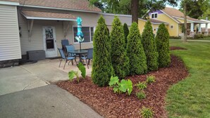 A residential house with a front porch, a small patio with chairs, and a well-maintained garden with trimmed bushes and a mulched area.