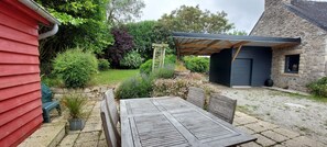 Un patio con mesa y sillas de madera, rodeado de un jardín con diversas plantas y un muro de piedra.