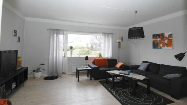 Una sala moderna con un sofá negro, una mesa de centro y un televisor sobre un mueble bajo. Hay una ventana con cortinas, una lámpara y una obra de arte en la pared.