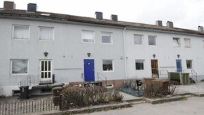 Una hilera de casas terreras blancas con ventanas y puertas uniformes, un pequeño jardín con una bicicleta y una valla metálica.