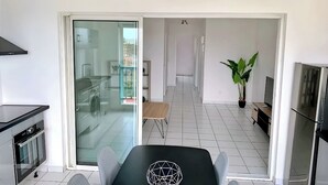 A modern kitchen with a built-in oven, a dining area with a table and chairs, a sliding glass door leading to a balcony, and a large potted plant. - Apparemment F2 vue mer (Les Trois-Îlets)