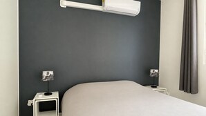 A modern hotel room with a bed, a wall-mounted air conditioner, a bedside table with a lamp, and a small picture frame. - Apparemment F2 vue mer (Les Trois-Îlets)