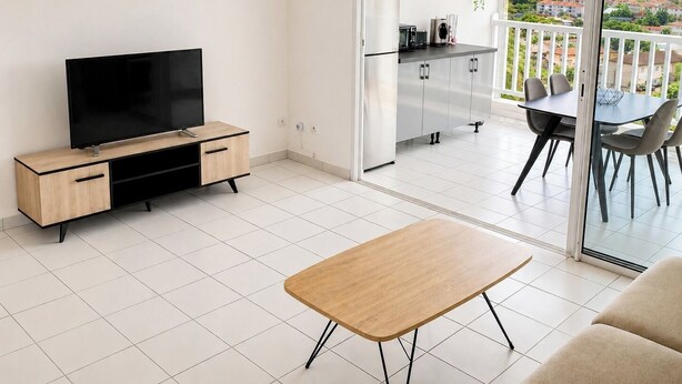 A modern living room with a flat-screen TV, a wooden coffee table, and a balcony with a dining area. - Apparemment F2 vue mer (Les Trois-Îlets)