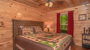 A wooden cabin bedroom with a bed, a nightstand, a window with red curtains, and a ceiling fan.