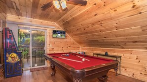 A wooden-paneled room with a pool table, a billiards table, and a Pac-Man arcade machine.