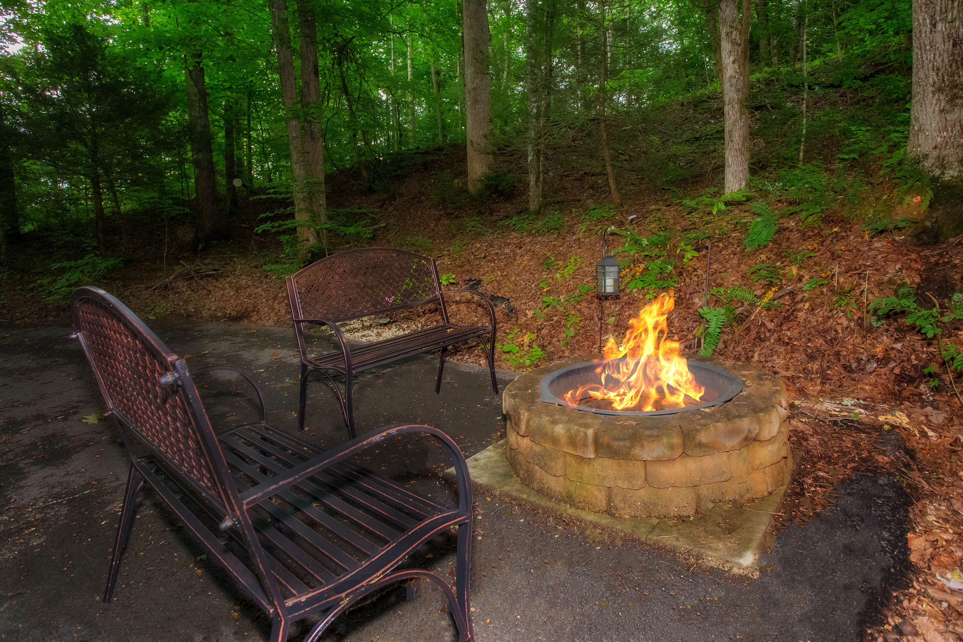 A fire pit with a metal bench and a metal chair in a forested area.