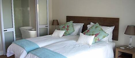 A double bed with floral pillows, a bedside table with a lamp, and a bathroom with a glass shower enclosure.
