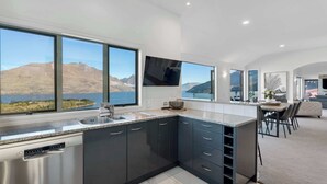 A modern kitchen with a large island, a dining area with a view, and a flat-screen TV mounted on the wall. - Million Dollar Views | Central Luxury | Private Hot Tub (Queenstown)